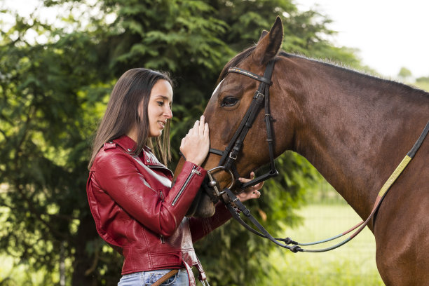 这位年轻女子在骑上马之前，一边看着它，一边抚摸着它的头。女子之前练习骑马。一个女人正准备在乡间小路上骑马图片下载