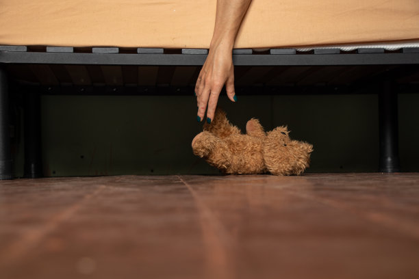 手靠近床在房子的木地板和床下躺着一只泰迪熊，家和舒适，找一只熊床下与你的手图片下载