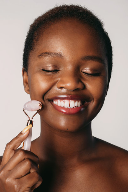拥有完美皮肤的非洲裔美国女人用玉辊按摩她的脸，在白色背景上闭着眼睛。自然美景。美丽的黑人女性。美丽的脸图片下载