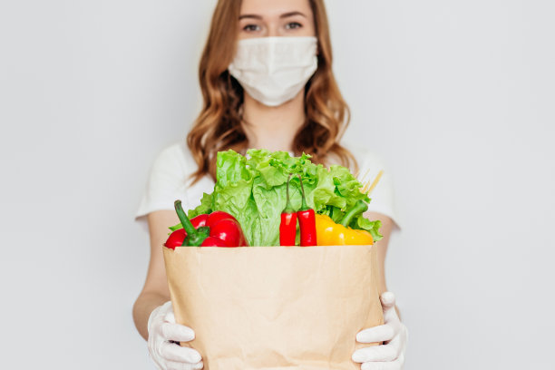 年轻的女快递志愿者戴着医用口罩，拿着一个纸袋，里面装着产品、蔬菜、草药，在白色、灰色的背景下隔离，隔离，冠状病毒，安全的食品配送图片下载