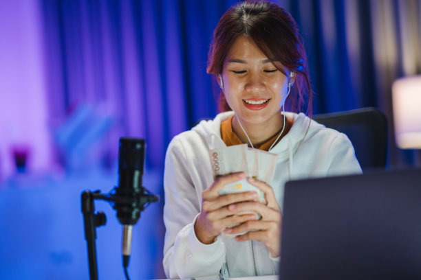 年轻的亚洲妇女拿着钱银行票据和录制播客在她的笔记本电脑在家庭工作室在晚上。图片下载