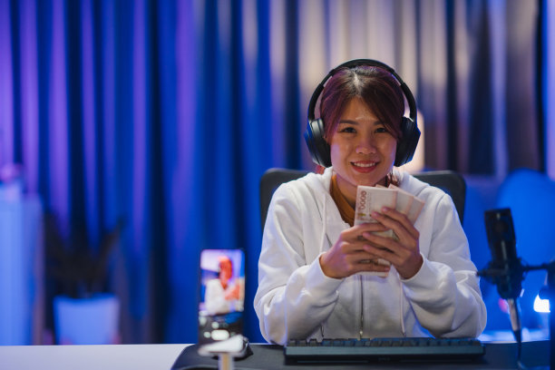 年轻的亚洲女性拿着钱，银行票据，玩在线电脑游戏，在家里的霓虹灯客厅里直播。图片下载