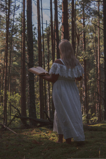 在森林里，一名女子穿着白色长裙，拿着一根棍子和一本书，背对着镜头站在那里。女巫站在那里，背对着森林的远处。万圣节图片下载