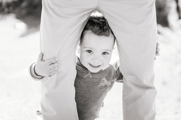 一个可爱的微笑的3岁男孩隐藏在一个成年人的腿后面，黑白相间图片下载