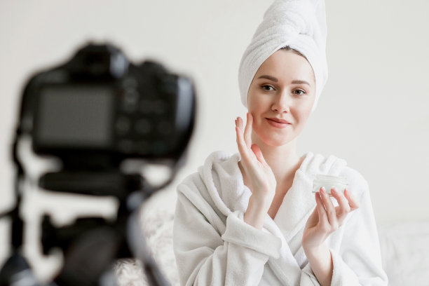 一名女博主身穿白大褂坐在床上，头上裹着毛巾，手里拿着面霜抹在脸上，一名年轻女子用摄像机录制了一段居家隔离期间的护理视频图片下载