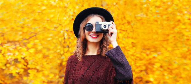 秋天肖像快乐微笑的年轻女子摄影师与胶卷相机戴圆帽，针织斗篷在公园的黄色树叶的背景图片下载