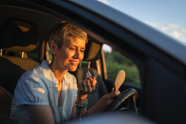 一个女人成熟的白人女商人坐在车里，涂口红，修妆，在她的脸上，等待在夏天的一天晚上真实的人复制空间灰色的头发图片下载