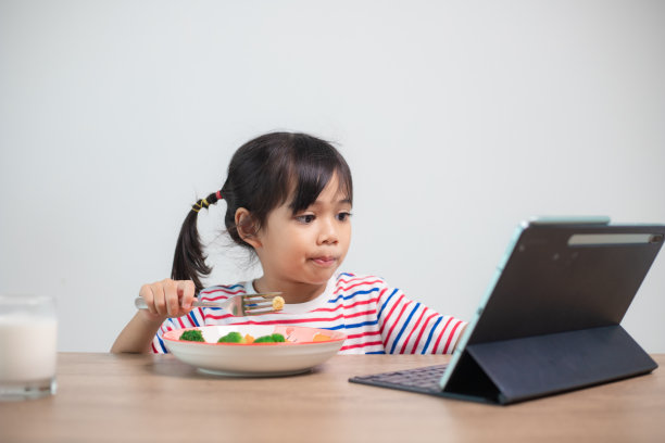 可爱的亚洲小女孩边吃午饭边看平板电脑上的电影。一个亚洲小孩正在吃晚饭，眼睛看着平板电脑上的卡通。全国饮食失调意识周。图片下载