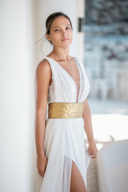 身穿白色无袖连衣裙、系着金色腰带的年轻美女靠在墙上，看着镜头图片下载