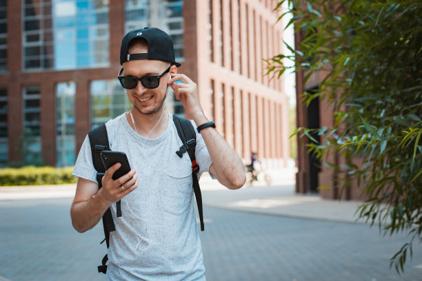 年轻人是一个时髦的家伙，戴着墨镜和黑色的帽子在城市里走，手里拿着智能手机，听着音乐耳机。T图片下载
