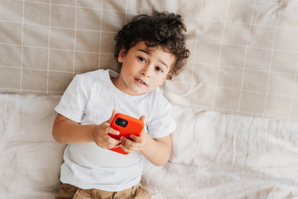 肖像从顶部英俊的卷发巴西卷发男孩拿着手机看相机躺在家里的床上。家庭休闲，孩子使用小玩意。帅气的白人小孩呆在家里。图片下载