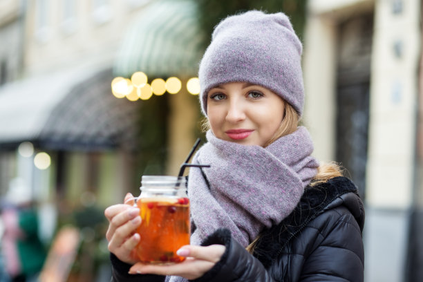 一个戴着围巾和帽子的年轻女孩正在街上喝热饮。走在圣诞大街上，手里捧着热茶。圣诞节销售图片下载