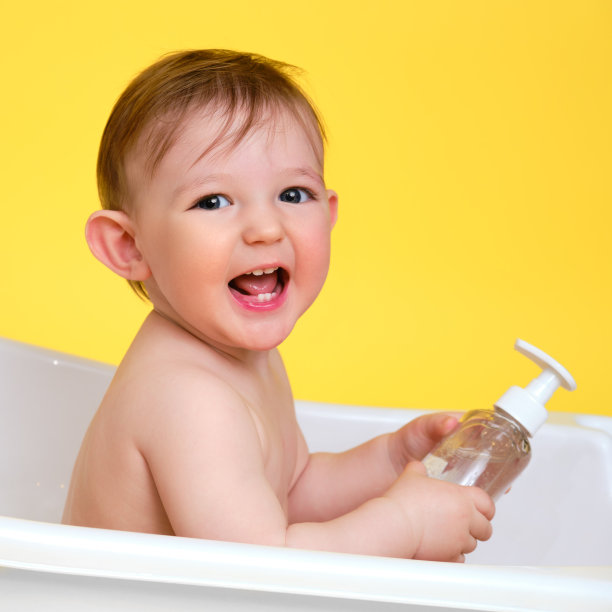 快乐的婴儿蹒跚学步的男孩与一瓶洗发水在白色浴缸上画室黄色的背景。一岁的微笑的孩子图片下载