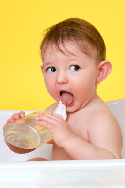 快乐的婴儿蹒跚学步的男孩与液体肥皂在一个白色浴缸在工作室的黄色背景。一岁的微笑的孩子图片下载