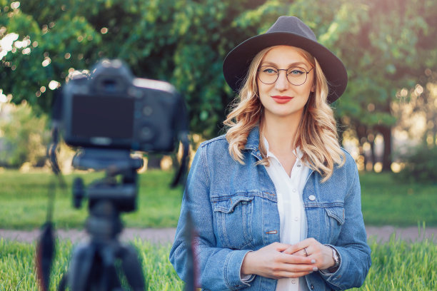 时髦的年轻女孩博主坐在街道上的草地上，拍摄vlog相机。年轻的金发女子作为博客录制视频教程图片下载