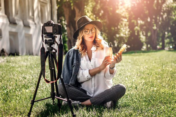 怀孕的女孩博主坐在街上的草地上，用相机拍摄vlog。女博主从太阳对宇宙手段进行了评论。年轻的金发女子作为博客录制视频教程图片下载