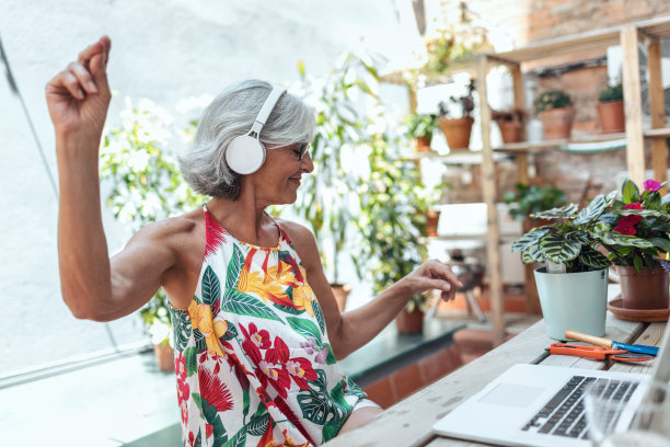 快乐的老妇人坐在温室里听音乐，用笔记本电脑。图片下载