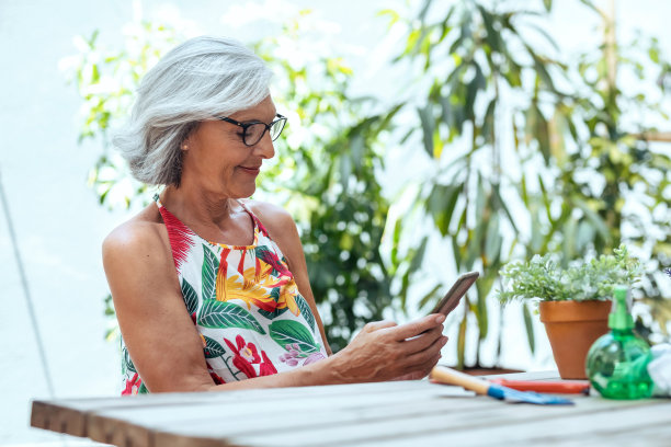 快乐的老年妇女在家里用智能手机拍绿色植物和鲜花。图片下载