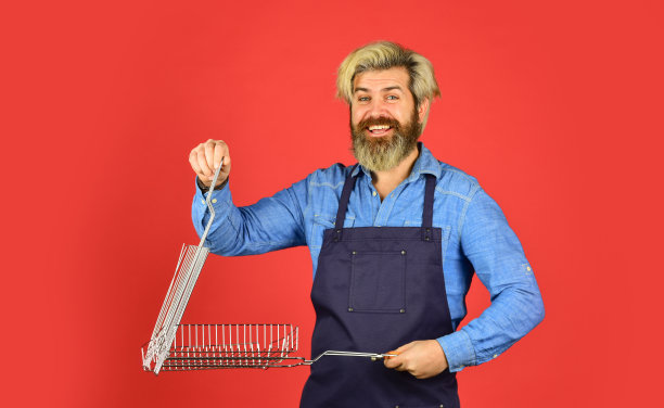 享受烹饪的乐趣。时髦的染胡子宣传烧烤设备。烹饪健康。烤肉的工具。穿着围裙的男人拿着烧烤架。烧烤食物。烹饪概念。烹饪用具。夏季野餐图片下载