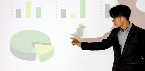 年轻的亚洲商人穿着西装，用投影仪屏幕做演示。带有饼状图和图表的白墙。培训员工、会议、报告、商学院。图片下载
