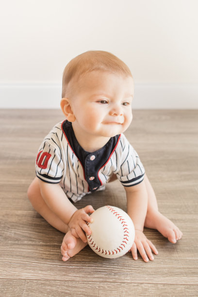 7个月大的小男孩穿着棒球服，一边玩垒球，一边学习一种新的发展技能，在家里的木地板上自己坐起来图片下载