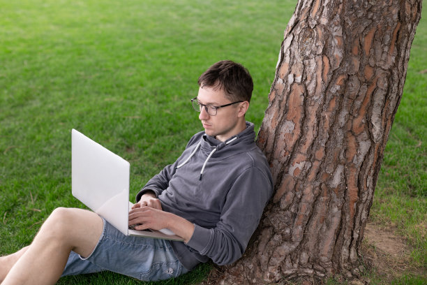 一个年轻人在草地上靠树坐着，看着他的笔记本电脑。他正在放松，享受着树影，在阳光明媚的日子里工作。图片下载
