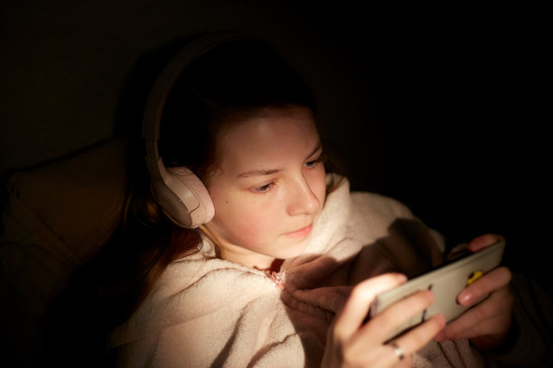 一个十几岁的女孩晚上在床上戴着无线耳机，手里拿着智能手机听流媒体音乐图片下载