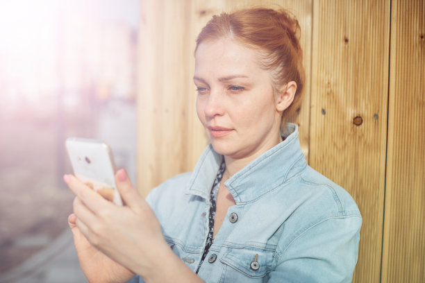 年轻的红发女子拿着手机坐在公园的长椅上，用移动设备在社交网络上交流图片下载