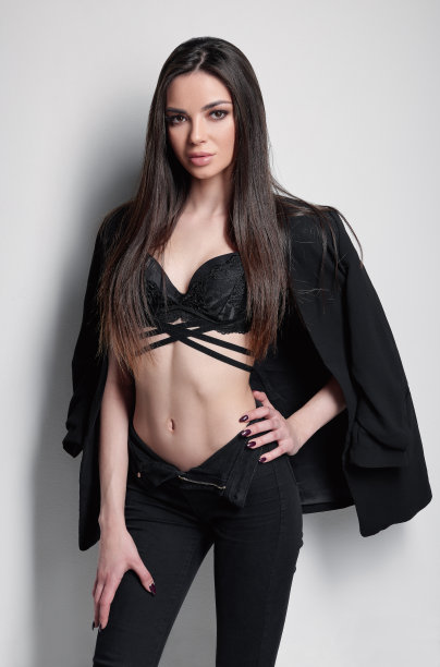 工作室时尚拍摄可爱的年轻女子。漂亮女孩的肖像穿着黑色夹克，牛仔裤和胸罩。靠在白墙上的性感女郎图片下载