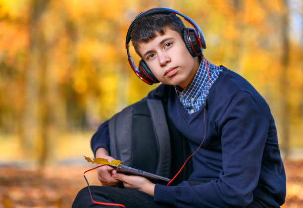 一个十几岁的男孩坐在秋天的公园里，戴着耳机听音乐，阳光明媚的日子里，树上的叶子都泛黄了图片下载
