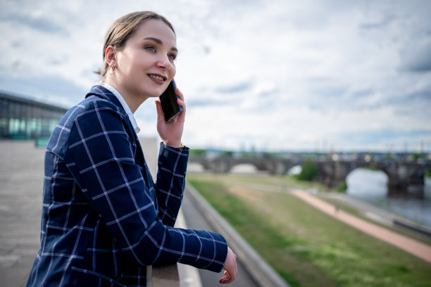 阴天的堤坝上，身穿格子夹克的女企业家斜靠在栏杆上，一边微笑一边讲手机图片下载