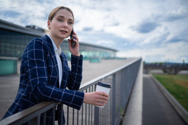 阴天的堤坝上，身穿格子夹克的女企业家斜靠在栏杆上，一边微笑一边讲手机图片下载