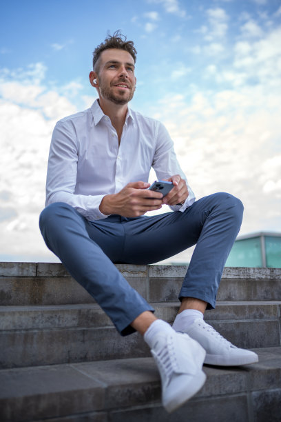 商人穿着正装，带着智能手机，坐在楼梯上听着音乐，面对着多云的蓝天，在街上休息图片下载