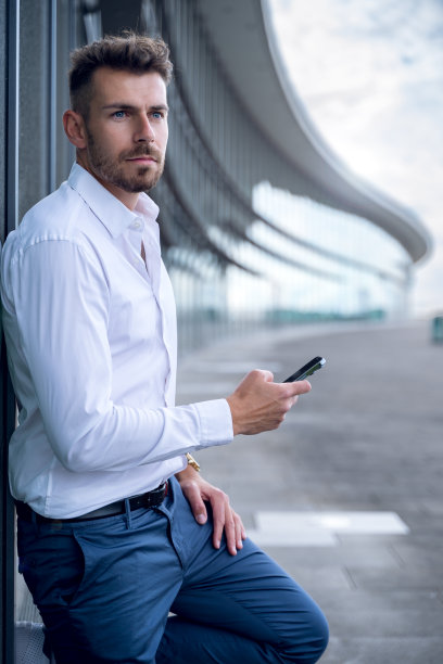 穿着正装的男性企业家倚在现代建筑的墙上，一边浏览智能手机，一边看向别处图片下载