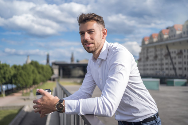 体贴的男经理穿着白衬衫，端着一杯咖啡，倚着栏杆，望着路堤上多云的蓝天图片下载