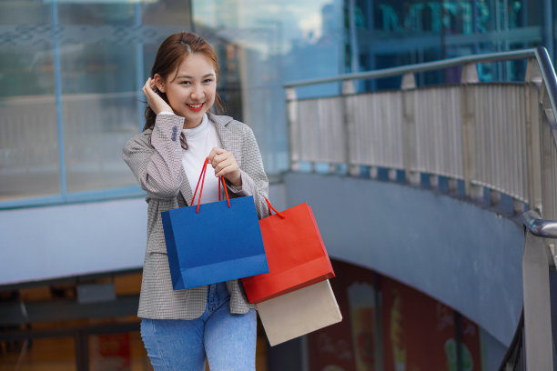 在购物中心前，一位拿着五颜六色的购物袋的亚洲女性正对着自己的名字微笑。图片下载