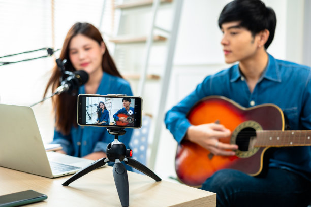 亚洲吉他手直播虚拟视频教程在家里，艺术家音乐家博主坐在沙发上在客厅。快乐青年歌手用乐器，在手机上作曲。图片下载