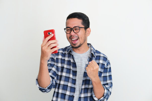 一名年轻的亚洲男子一边看手机，一边握紧拳头表示兴奋图片下载