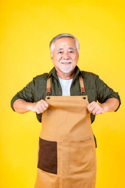 肖像迷人的亚洲退休老人有白色的小胡子，穿着围裙的胡子指向黄色的围裙背景。图片下载
