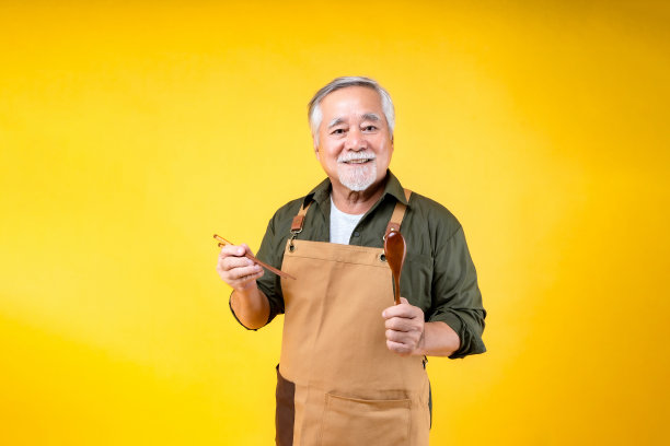 肖像迷人的退休亚洲老人有白色的胡子和胡子穿着围裙拿着叉子勺子黄色的背景。图片下载