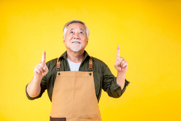 肖像迷人的亚洲退休老人留着白色的小胡子，戴着围裙指着胡子，双手临摹空间。图片下载