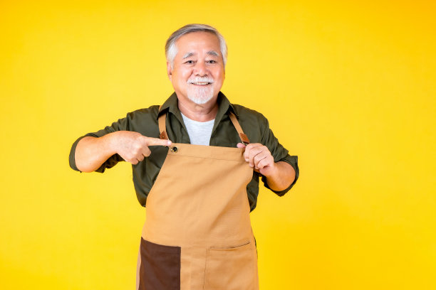 肖像迷人的亚洲退休老人有白色的小胡子，穿着围裙的胡子指向黄色的围裙背景。图片下载