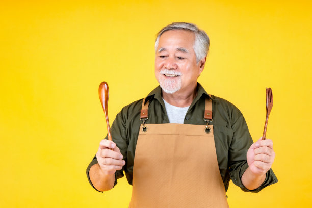 肖像迷人的退休亚洲老人有白色的胡子和胡子穿着围裙拿着叉子勺子黄色的背景。图片下载