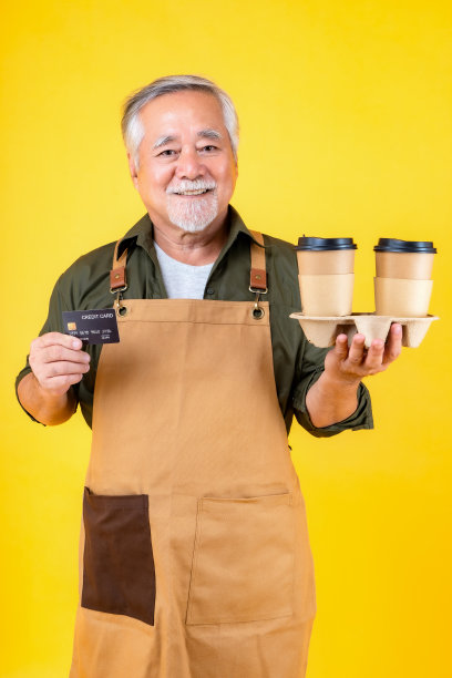肖像迷人的退休亚洲老人有白色的胡子和胡子，穿着围裙拿着纸杯咖啡和信用卡。图片下载