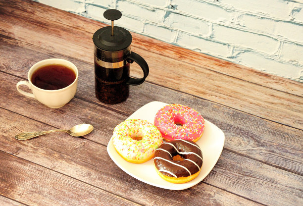 木桌上摆着三块多彩釉甜甜圈的盘子，汤勺一杯热茶，茶壶一个。图片下载