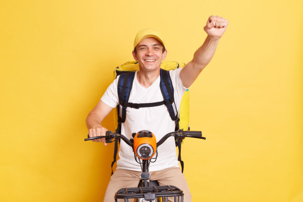 微笑，快乐的快递员戴着帽子，带着热背包，在电动滑板车上举起拳头，快速送餐，表达积极的情绪，孤立在黄色的背景上。图片下载