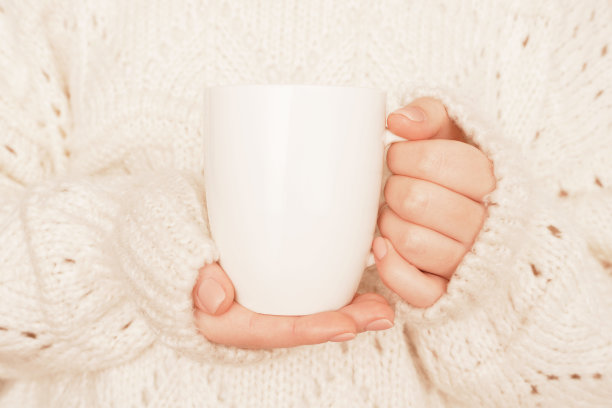 女人的手拿着模拟的白色杯子，杯子特写。女性穿着白色针织毛衣。空白模板，标志，文字设计，宣传，文字信息。冬天,秋天的概念。图片下载