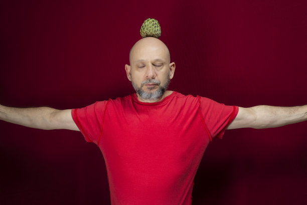 年轻英俊的男子与胡子，光头和红衬衫站与松果平衡的头在红色的背景。图片下载