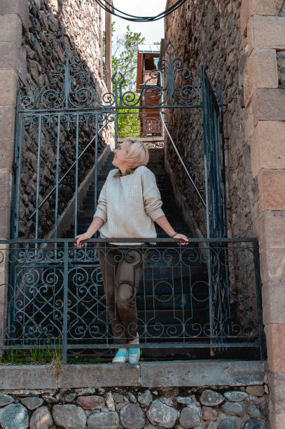 身穿夹克和裤子、脚穿白色运动鞋的年轻金发女子双手扶在两座石屋之间的栏杆上图片下载