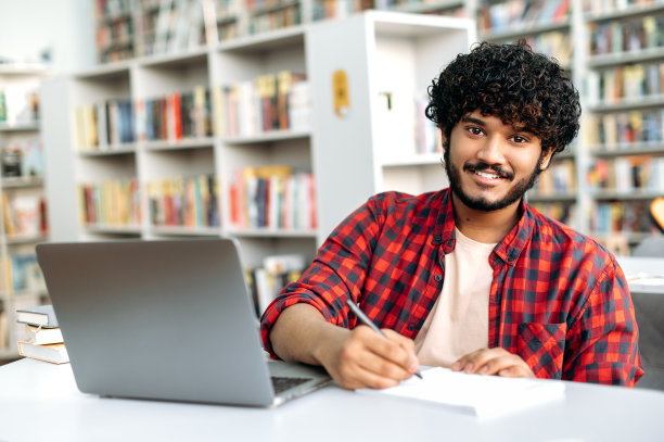 一个英俊积极的学生或自由职业者的照片，阿拉伯或印度裔卷发，坐在图书馆的桌子上，用笔记本电脑做笔记，看着相机，微笑图片下载